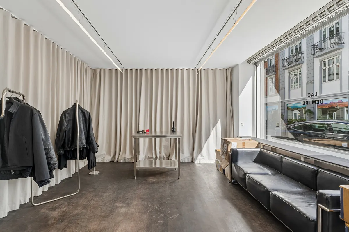 En moderne butikslokale med store hvide gardiner og trægulv, udstyret med en lædersofa og en stålbord, og udsigt til en parkeret bil og bygninger med balkoner