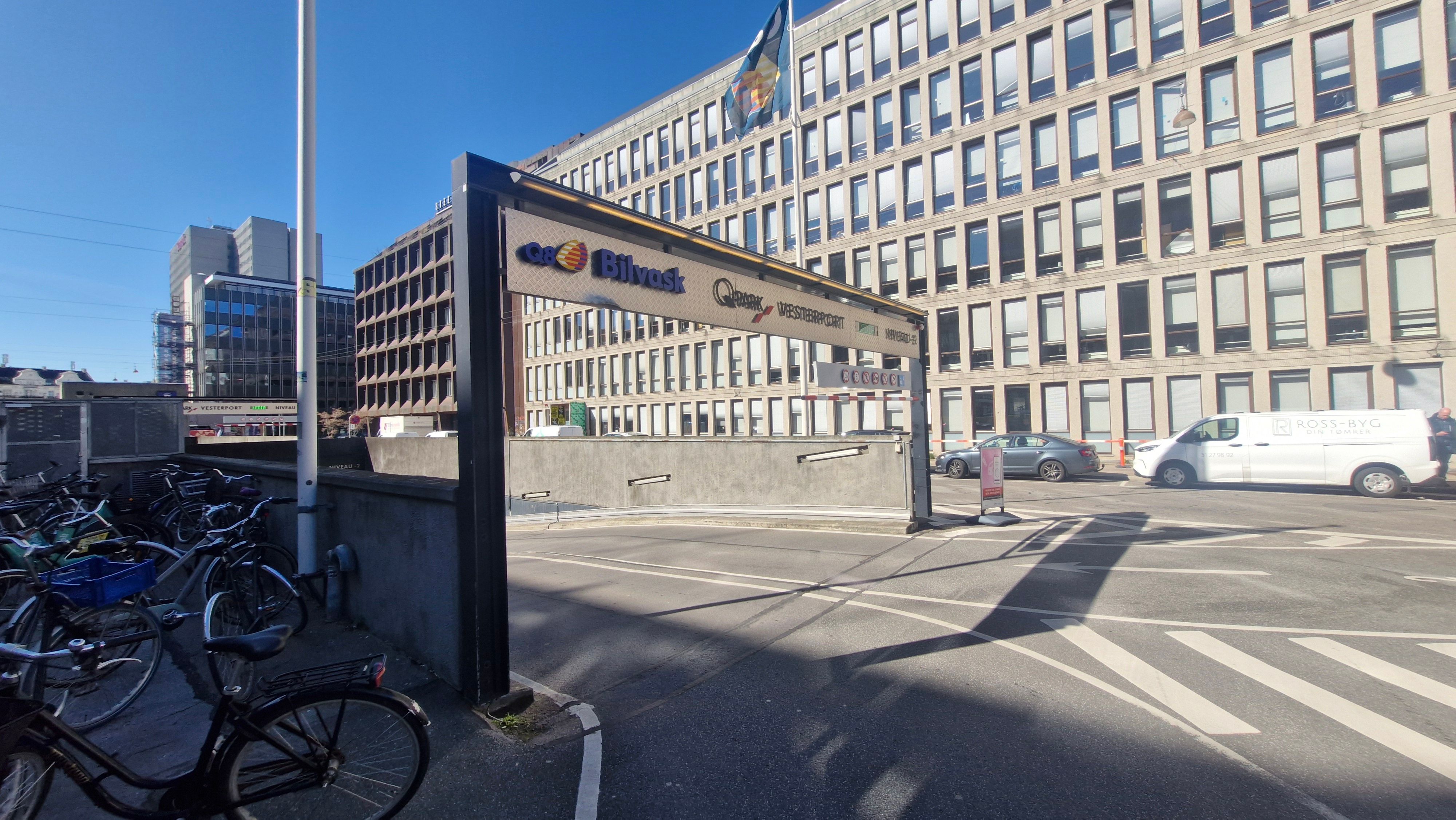 Flere cykler parkeret ved en stor bygning med skiltet Bilvask og Q8 West. En bilparkeringsplads med biler og en person i baggrunden.