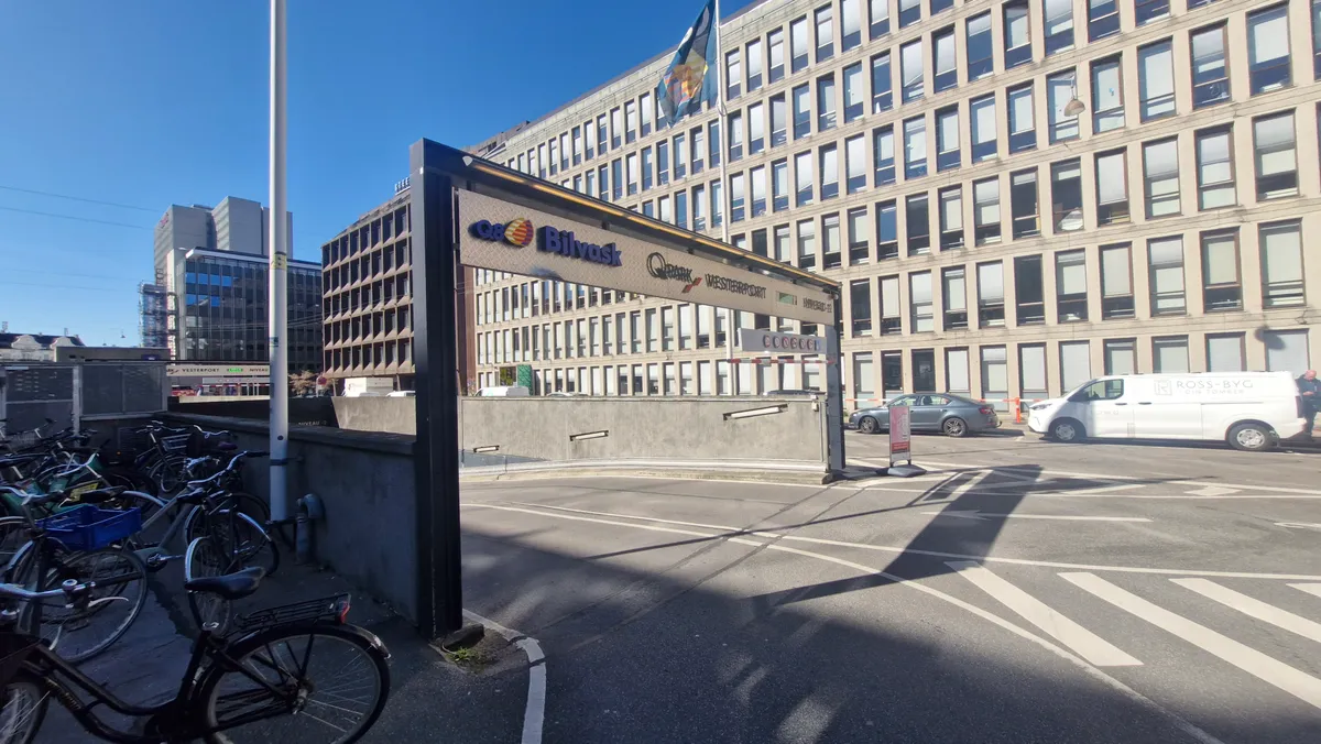 Flere cykler parkeret ved en stor bygning med skiltet Bilvask og Q8 West. En bilparkeringsplads med biler og en person i baggrunden.