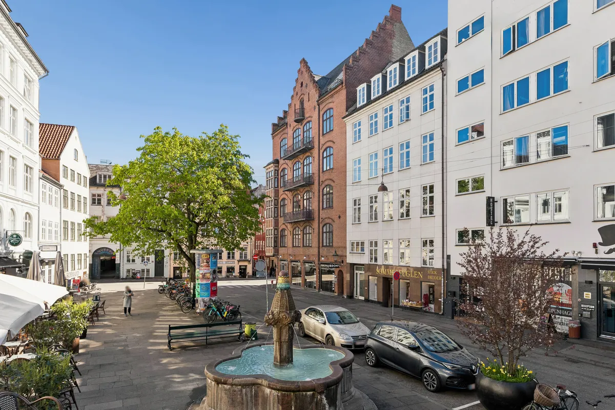 Lyst kontor i Vandkunsten 3 i København K med vandfontæne og grønne planter foran. Parkerede biler og gågade med butikker og caféer.