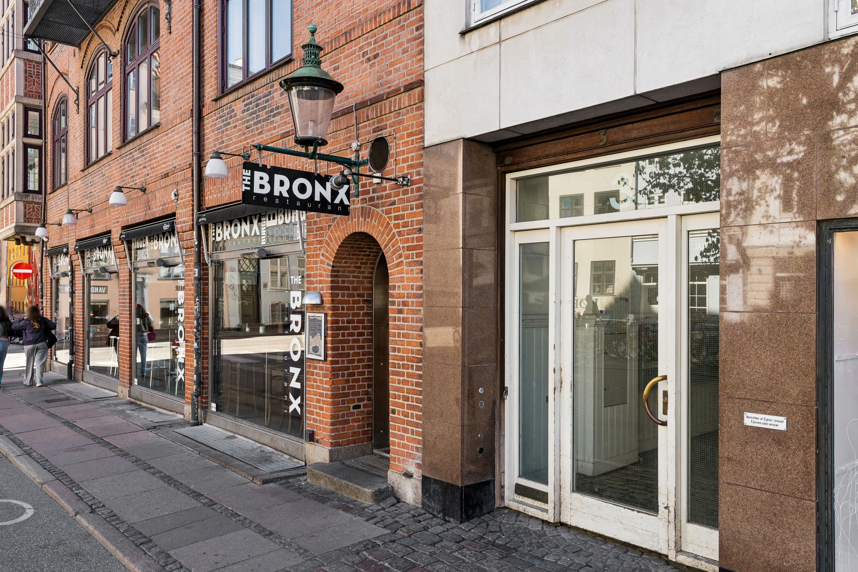 Lyst kontor i røde mursten med The Bronx skilt og glasdøre med håndtag, sammen med personer, der går forbi udenfor
