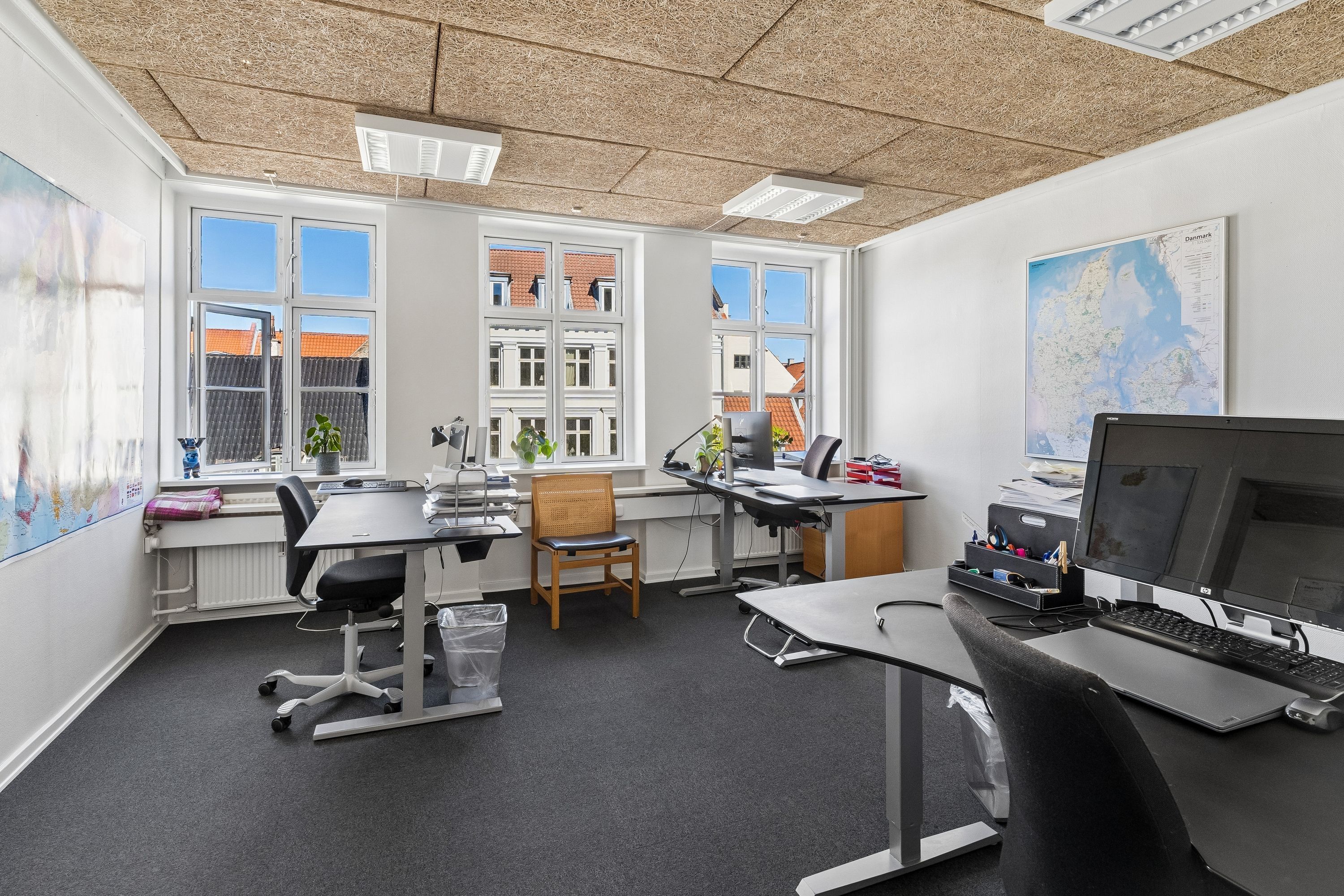 Kontor til leje i København K med lyse kontorer, moderne møbler og udsigt til byen. Kort, plante og skrivebord med computer og skrivebordslampe.