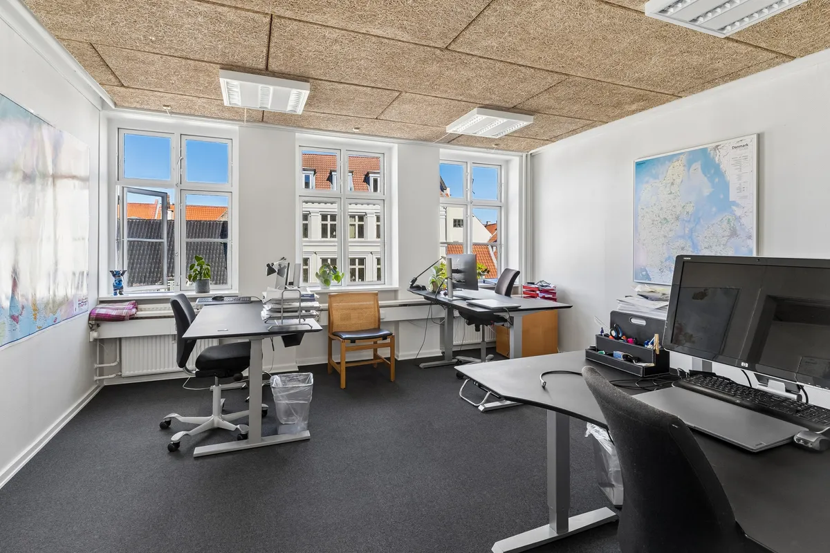 Kontor til leje i København K med lyse kontorer, moderne møbler og udsigt til byen. Kort, plante og skrivebord med computer og skrivebordslampe.
