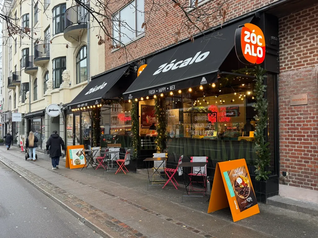Restaurant med navnet Zocalo i en gade med folk, der går forbi og bord med stole og træstole samt træborde og stolpe