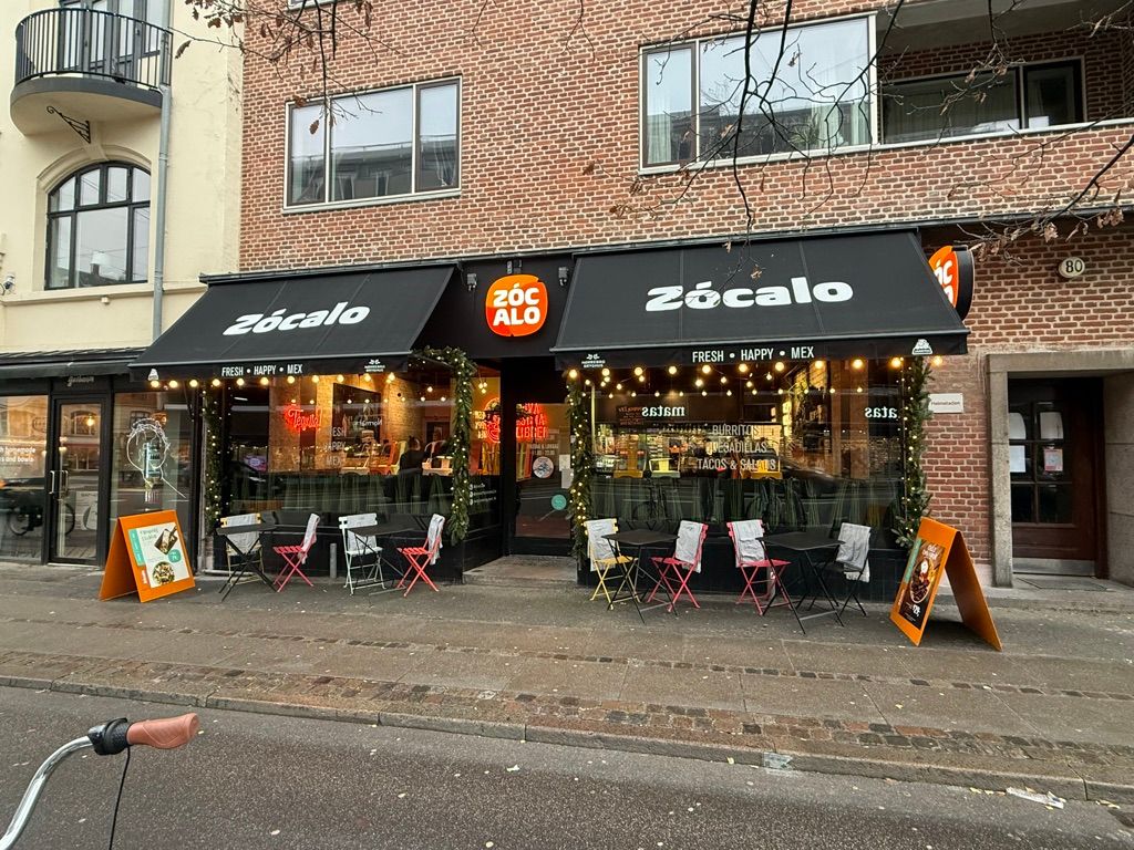 Restaurant med udendørsborde og stole, træplanter og juledekor, træklap med gult logo og orange skilt, gulvbelægning, cykel og røde træstole