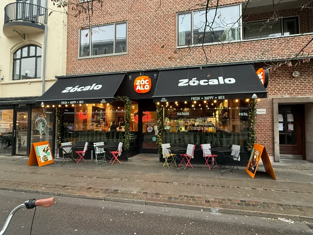 Restaurant med udendørsborde og stole, træplanter og juledekor, træklap med gult logo og orange skilt, gulvbelægning, cykel og røde træstole