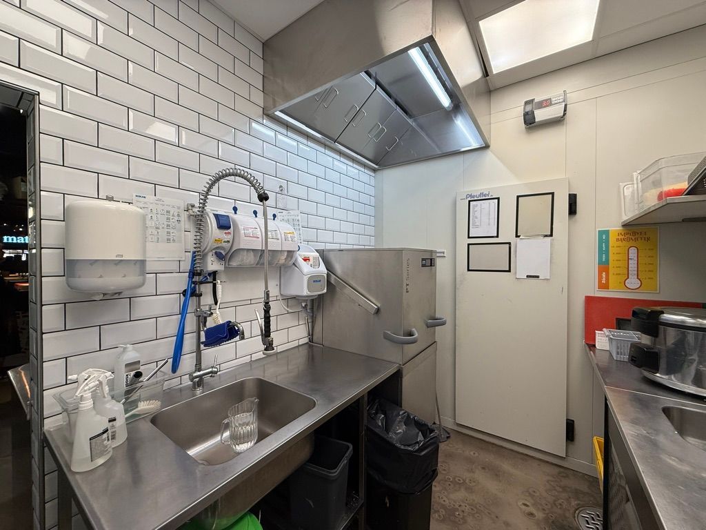 Røgsuger og ventilation i kombineret køkken og vaskeområde. Væggene er klædt med hvide fliser og der er en dør i højre side. Der er et glas og rengøringsmidler i vasken og der er to skraldespande.