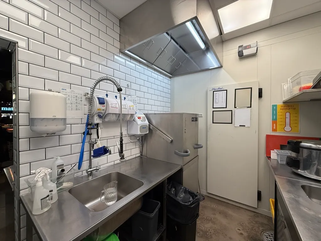 Røgsuger og ventilation i kombineret køkken og vaskeområde. Væggene er klædt med hvide fliser og der er en dør i højre side. Der er et glas og rengøringsmidler i vasken og der er to skraldespande.