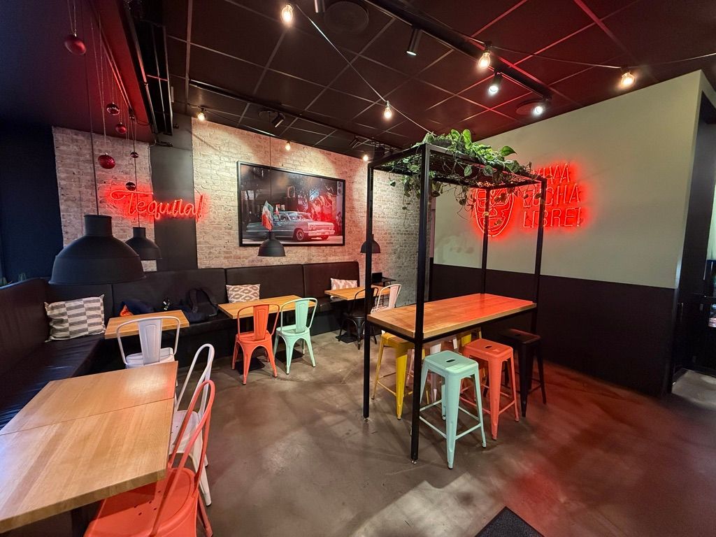 Et trendy restaurantlejemål med røde neonlygter og lysepunkter på loftet. Bord og stole samt sofaer med pudsede puder og plante i krydsfinerhylden