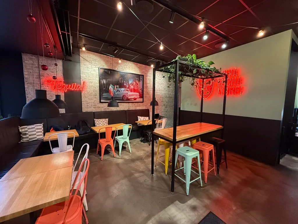 Et trendy restaurantlejemål med røde neonlygter og lysepunkter på loftet. Bord og stole samt sofaer med pudsede puder og plante i krydsfinerhylden