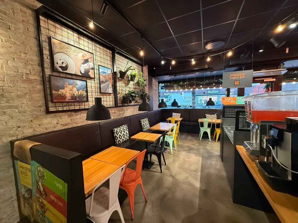 Interiøret i en restaurant med bord og stole, lysekroner, væg med malerier og plante, og en kaffemaskine ved siden af et vinduesvis.