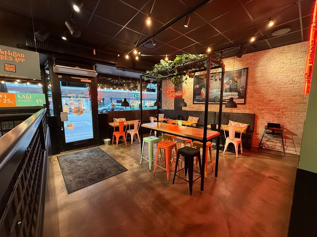 Indre af en restaurant med bord og stole samt en terrasse med et bord og stole. Der er en stor vinduesvisning og et vægmaleri med en bil. Der er et bord med en plante og et lys.