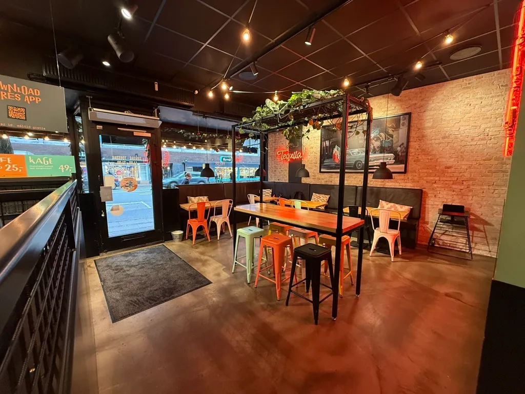 Indre af en restaurant med bord og stole samt en terrasse med et bord og stole. Der er en stor vinduesvisning og et vægmaleri med en bil. Der er et bord med en plante og et lys.