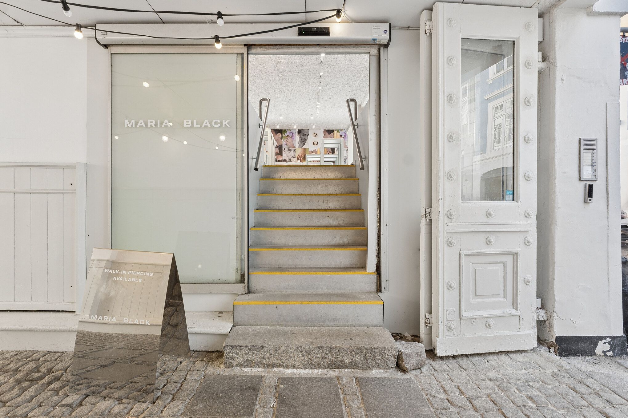 Stilren butik med gulgule trin og hvide vægge. Maria Black logo på glassdøren. På gulvet står en reflekterende plakat med teksten Walk-in Piercing Available.