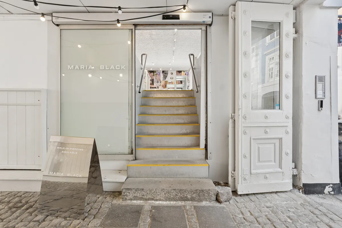 Stilren butik med gulgule trin og hvide vægge. Maria Black logo på glassdøren. På gulvet står en reflekterende plakat med teksten Walk-in Piercing Available.