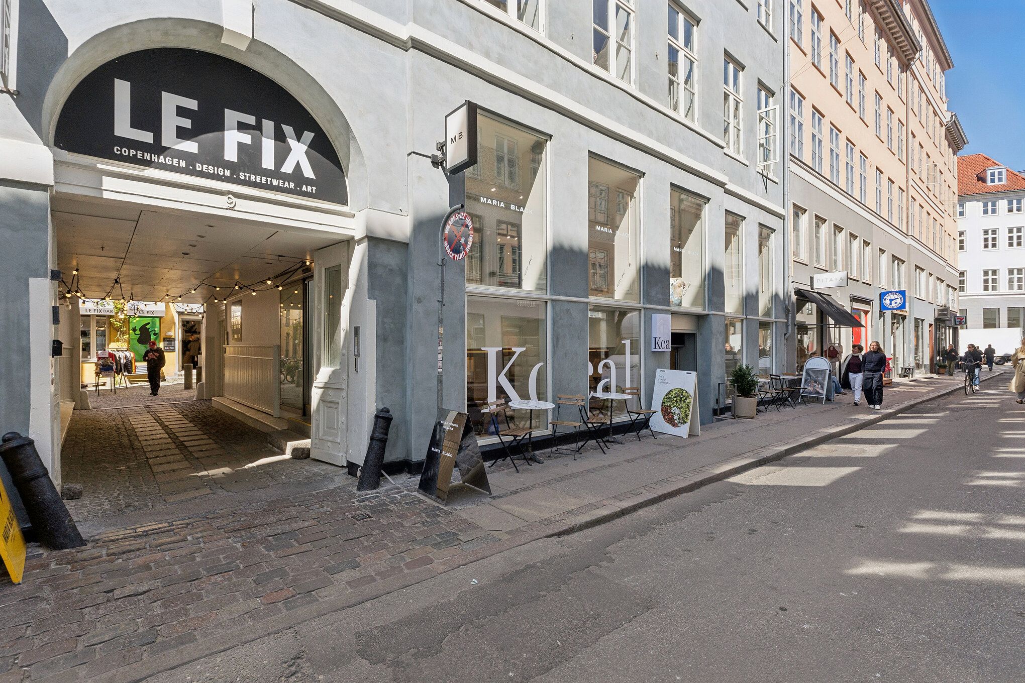 Le Fix-butik med design, streetwear og kunst i København. Gade med gavle og folk i baggrunden, bord og stole foran butikken og søjler med skilte