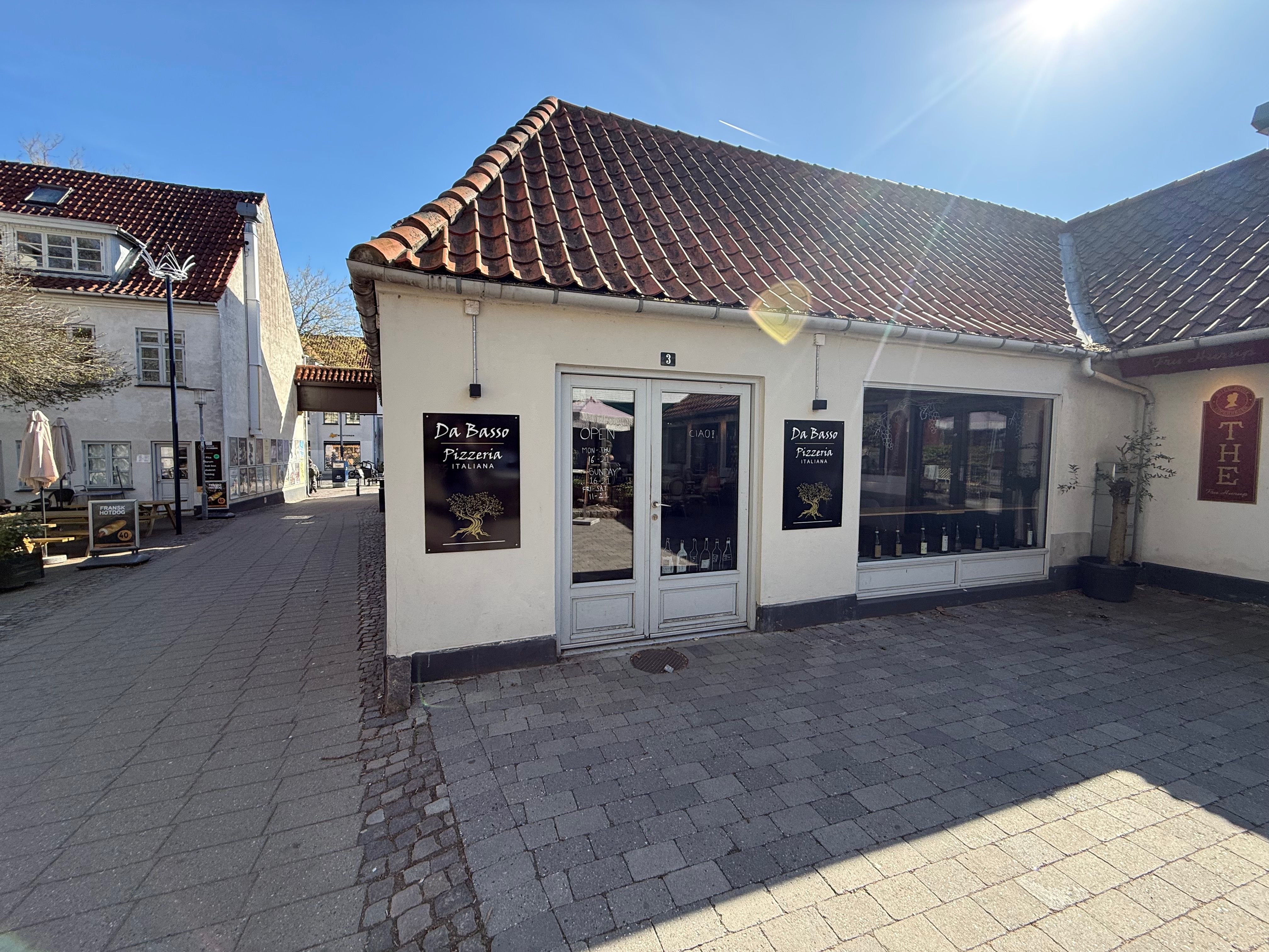 Take-away til leje i Kongens Lyngby. Opført i hvid mursten med to store vinduer og døre. Stående plante og to paraplyer. Nummer 3 på væggen.