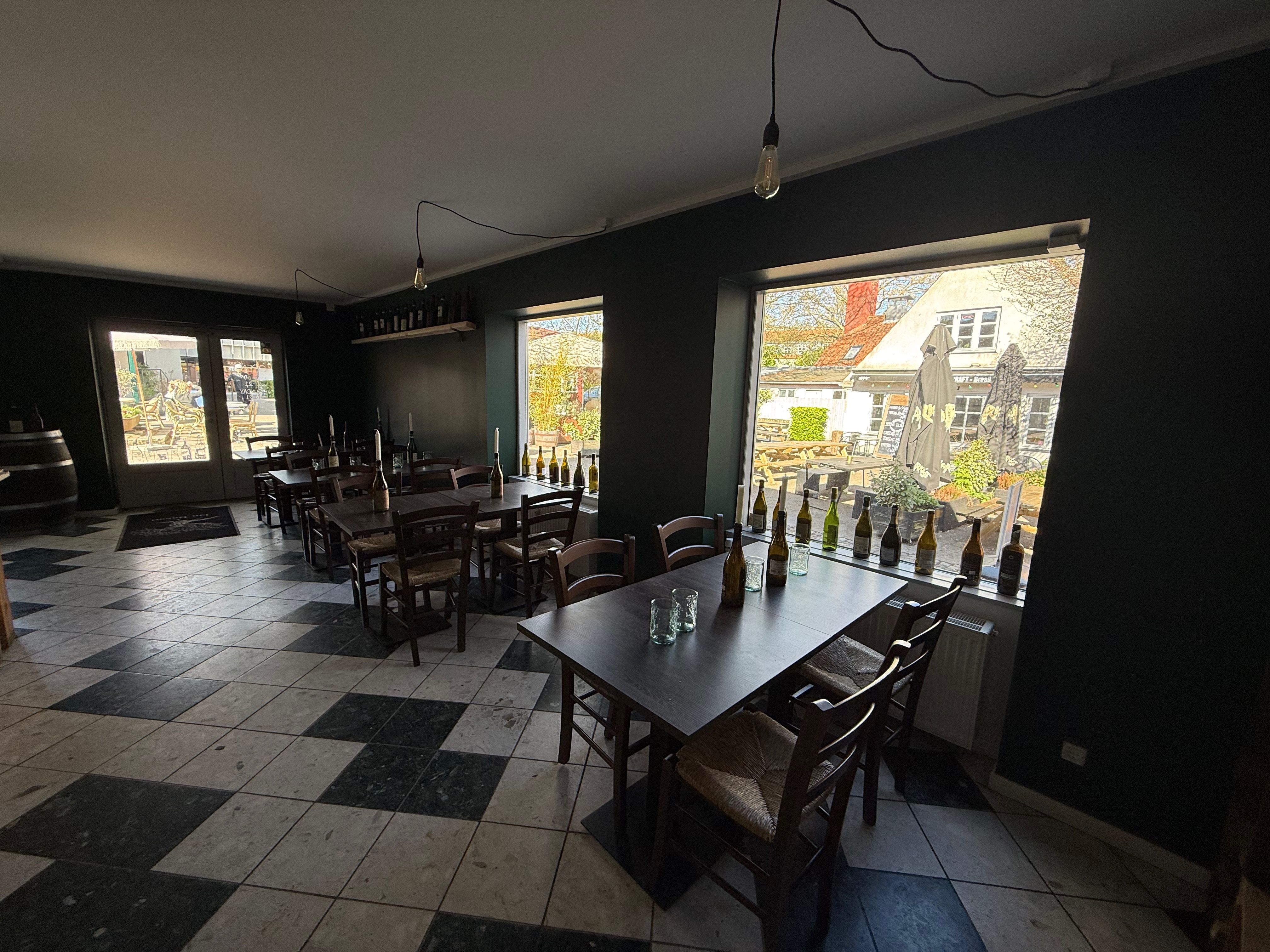 Take-away til leje på Likørstræde 3, 2800 Kongens Lyngby. Værelse med træborde og stole, vinduer med udsigt til byen, lyse pærelampetter og flere vinflasker