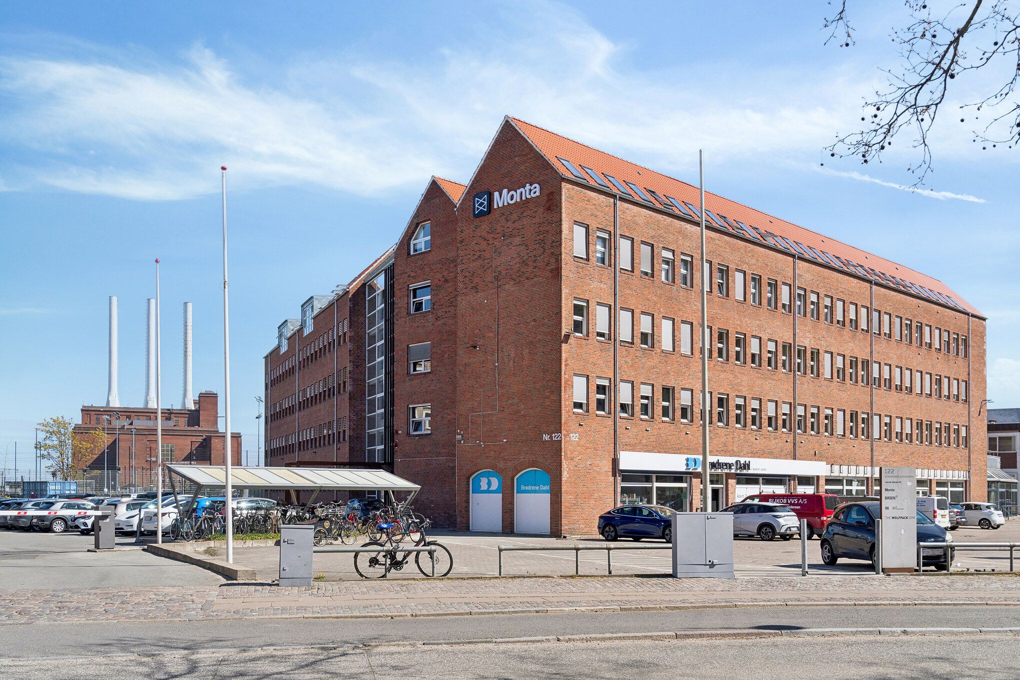 Monta kontorlejemål med mange parkeringspladser og flere biler parkeret. Foran bygningen er en bro og en bollard. Et enkelt cykelstativ med cykel. Adgang til kontoret er gennem to store hvide døre