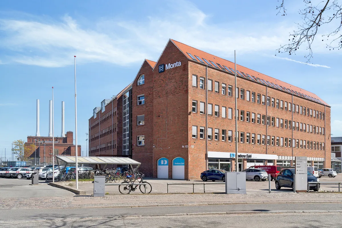 Monta kontorlejemål med mange parkeringspladser og flere biler parkeret. Foran bygningen er en bro og en bollard. Et enkelt cykelstativ med cykel. Adgang til kontoret er gennem to store hvide døre
