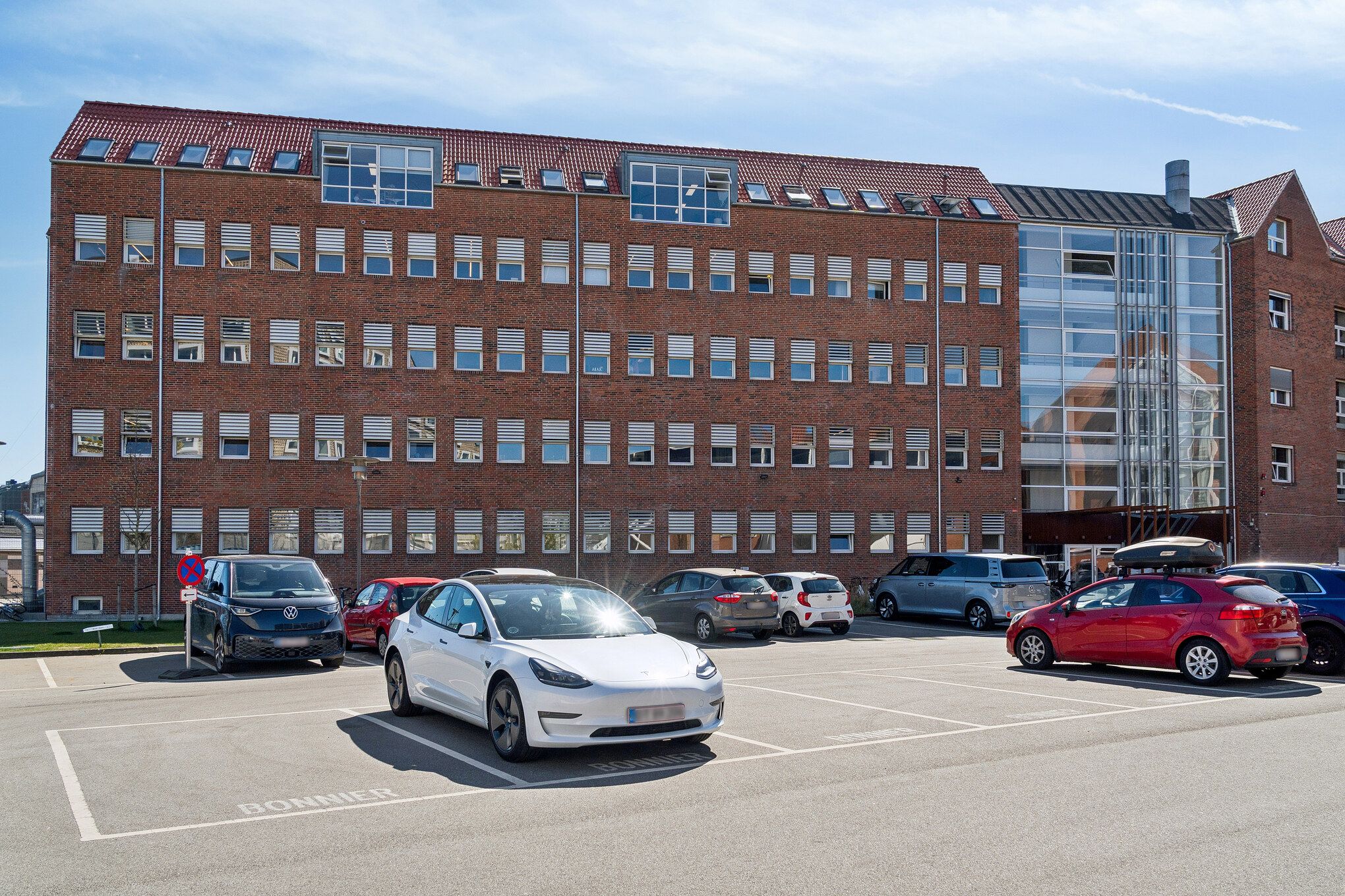 Parkerede biler, herunder en hvid Tesla Model 3, foran en stor rød murstensbygning med mange vinduer og en glastavle