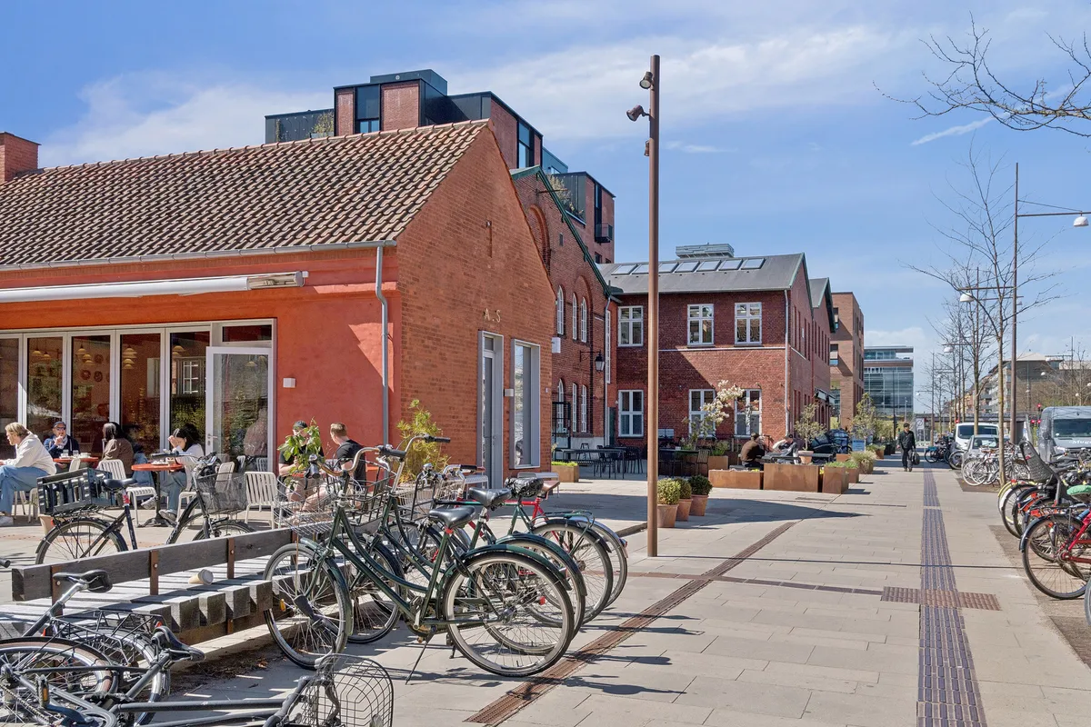 Mange cykler står parkeret på fortov og facaden af en rødmalet bygning med store ruder og bænke foran