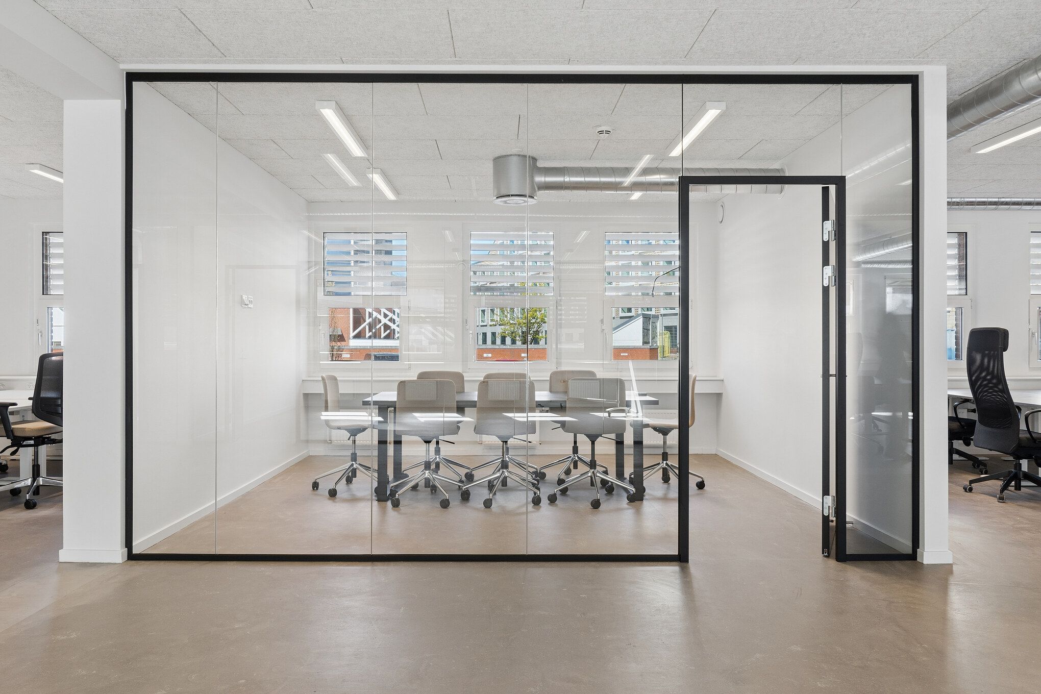 Glassvægge og højt loft med indbygget belysning. Kontorbord med stole og vinduer med skyggeskærme. Moderne indretning med hvid væg og gulv.