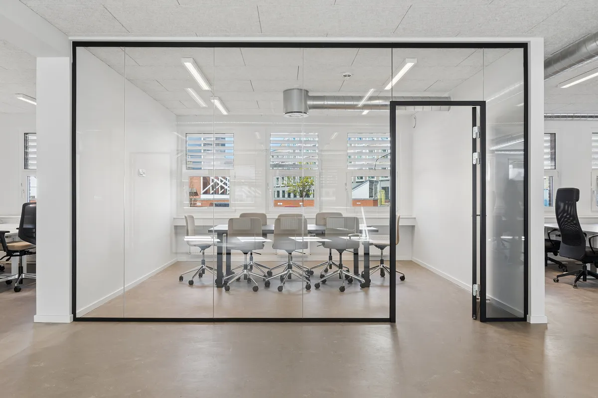 Glassvægge og højt loft med indbygget belysning. Kontorbord med stole og vinduer med skyggeskærme. Moderne indretning med hvid væg og gulv.