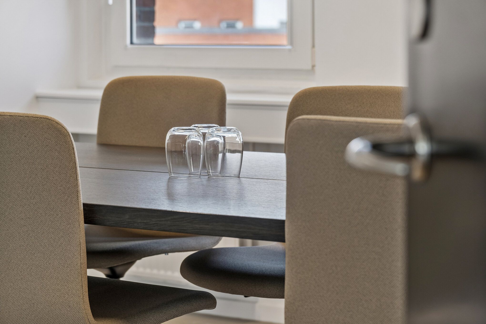 Moderne kontorrum med fire beige stole omkring et slankt bord med fire glas, tæt ved et vindue med en udsigt til byens bygninger
