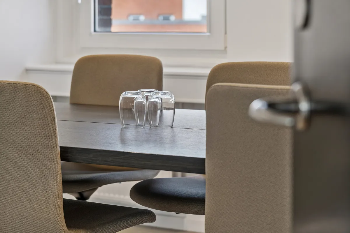 Moderne kontorrum med fire beige stole omkring et slankt bord med fire glas, tæt ved et vindue med en udsigt til byens bygninger