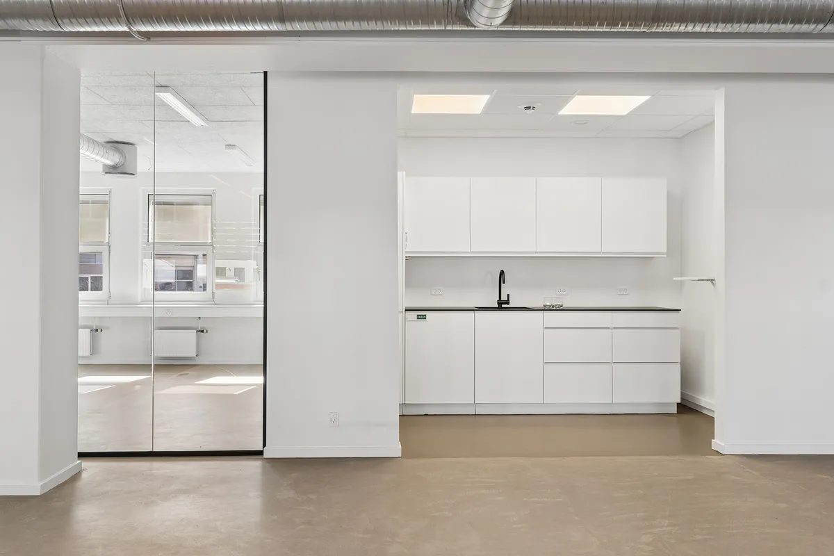 Minimalistisk kontor med hvide vægge, glasdøre, højloft og lyse linjerne i loftet. Hjørne med køkken, hvidt køkkenskab og rustfrit stål vandhane