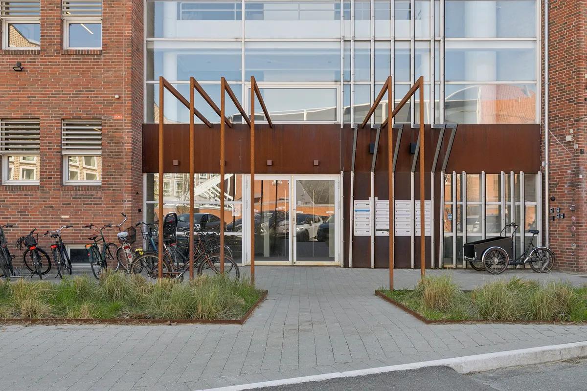 Moderne kontorlejemål med glasdøre og rustfri stålstruktur i forgrunden, parkerede cykler og et lastcykel med trailer, røde mursten og vinduer på baggrunden
