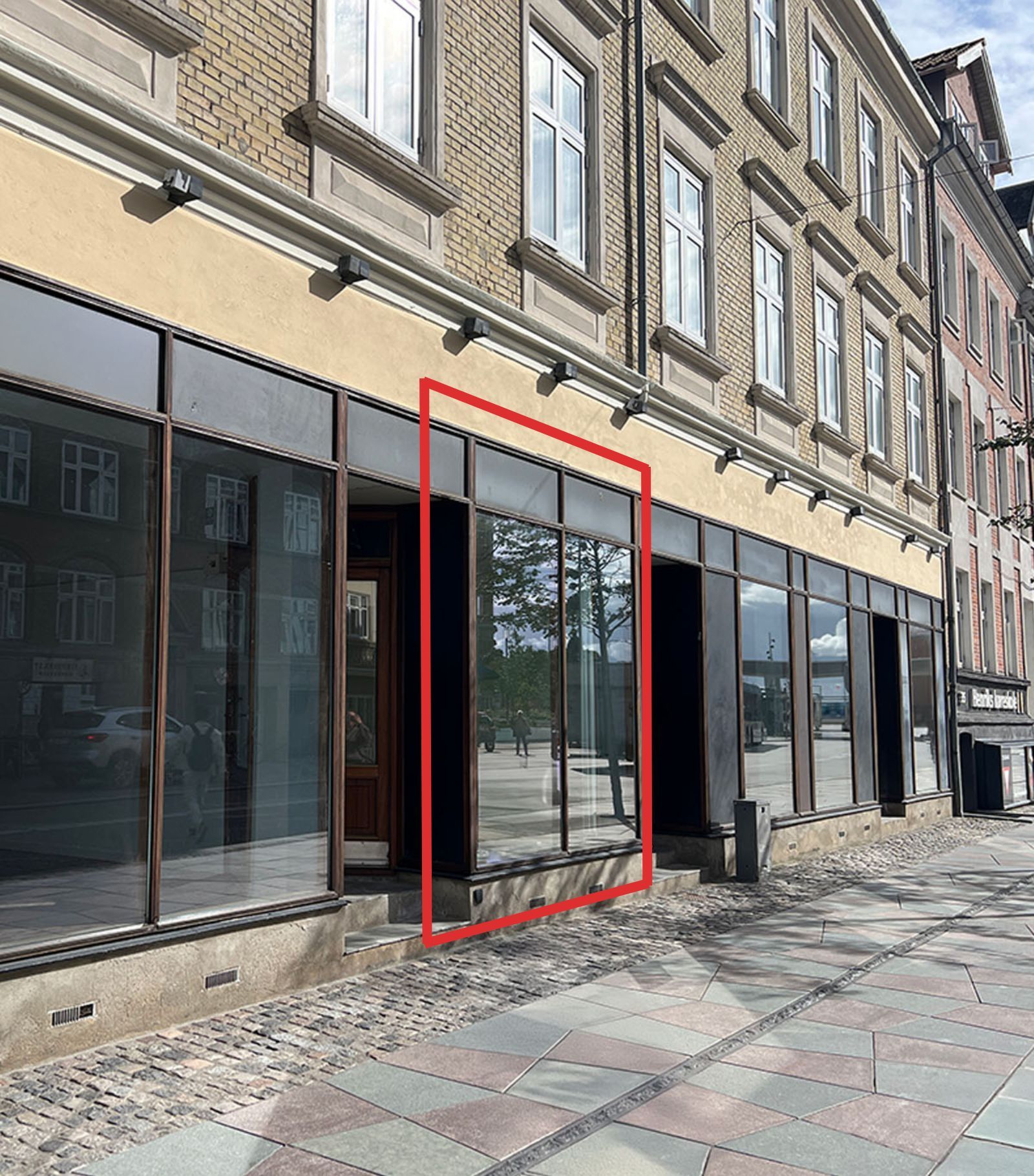 Butik med glasvinduer og en dør på Boulevarden 33B i Aalborg, udsmykket med gule og røde lys. Baggrund med røde linjer markerer vindue og dør.