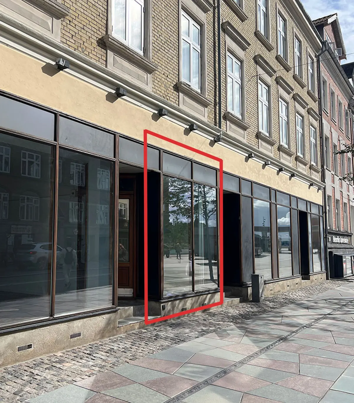 Butik med glasvinduer og en dør på Boulevarden 33B i Aalborg, udsmykket med gule og røde lys. Baggrund med røde linjer markerer vindue og dør.