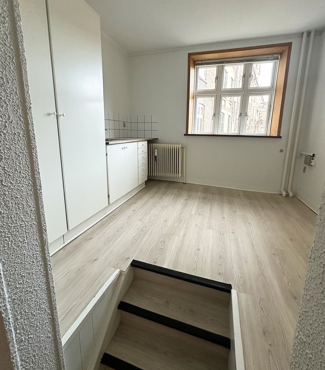Udstødende loft og hvide vægge. To hvide skabe og køkken med radiator. Gulv med lyse, træagtige bjælker. Trin nedad.