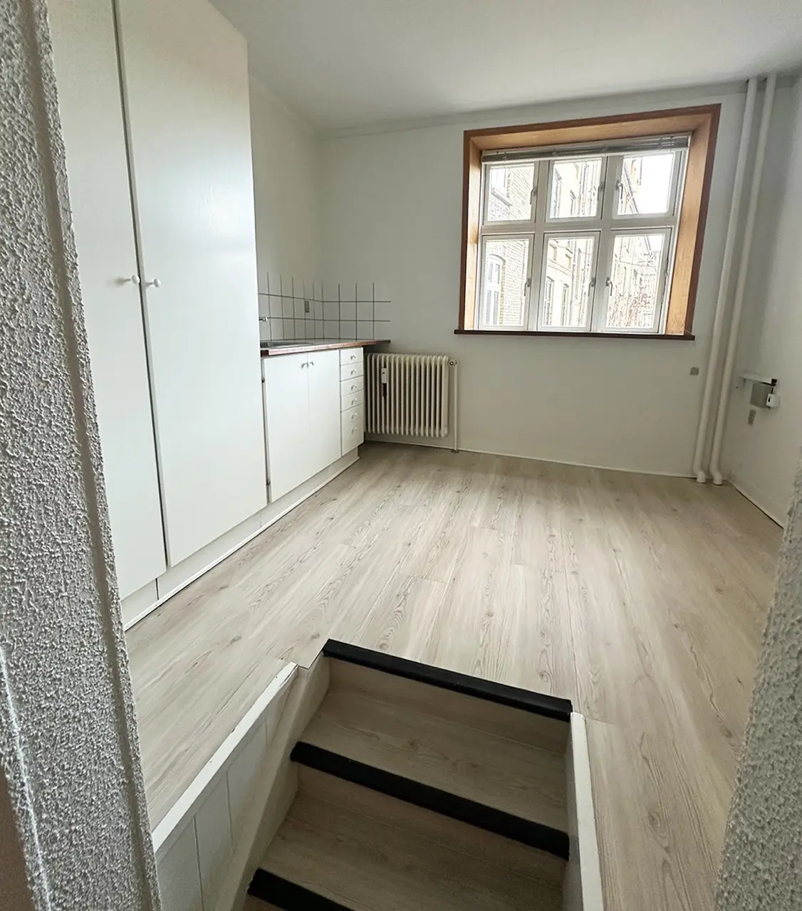 Udstødende loft og hvide vægge. To hvide skabe og køkken med radiator. Gulv med lyse, træagtige bjælker. Trin nedad.