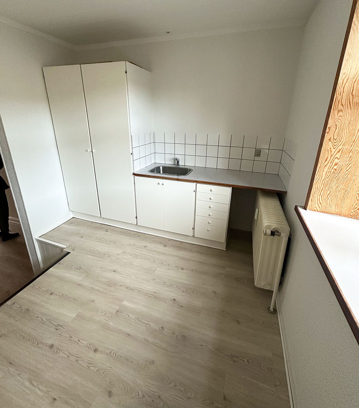 Lejligheds køkken med hvidt loft, gulv og vægge. Væggen med lysegrå klinker. Sort skab med 3 døre. Hvidt vasketøjsskab. Hvidt køkken med en hætte og et nyt radiator