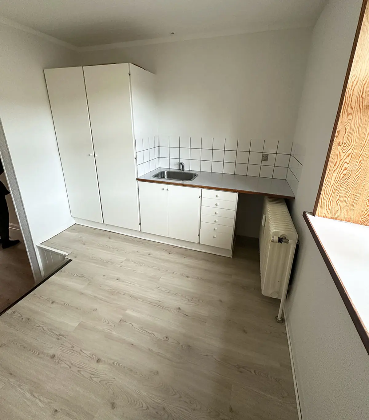 Lejligheds køkken med hvidt loft, gulv og vægge. Væggen med lysegrå klinker. Sort skab med 3 døre. Hvidt vasketøjsskab. Hvidt køkken med en hætte og et nyt radiator