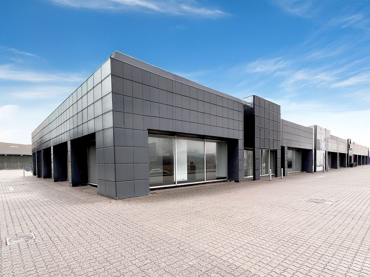 En moderne kommerciel bygning med store glasvinduer og en grøn tagkant er udsat til leje i Hjørring. Bygningen er lukket og har en lukket parkeringsplads med brolægning