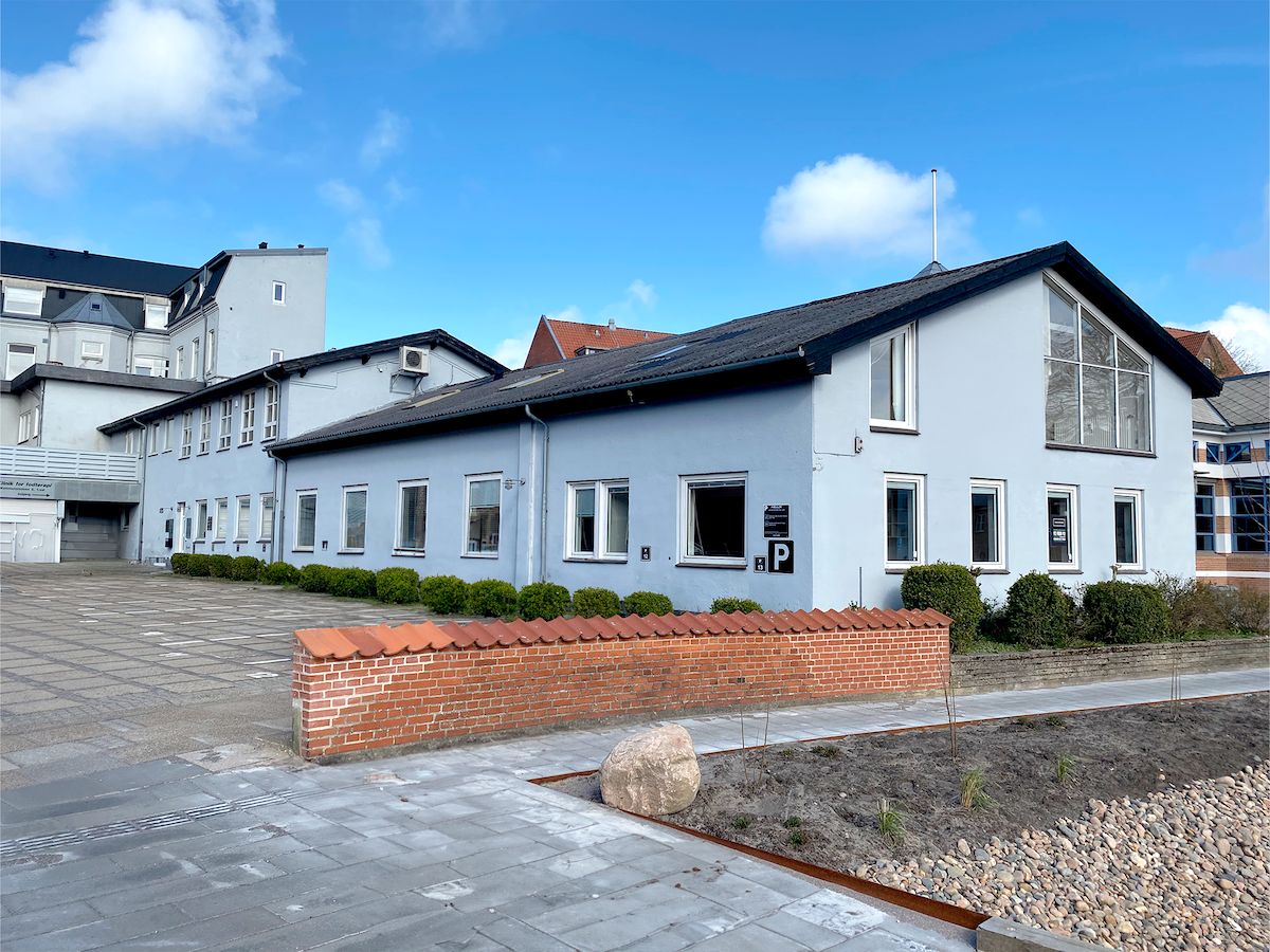 Moderne bygning med glasvinduer og sort tag på Mammutpladsen 5, 9800 Hjørring. Murstensvæg og grønne buske udenfor. Parkeringsskilt på facaden.