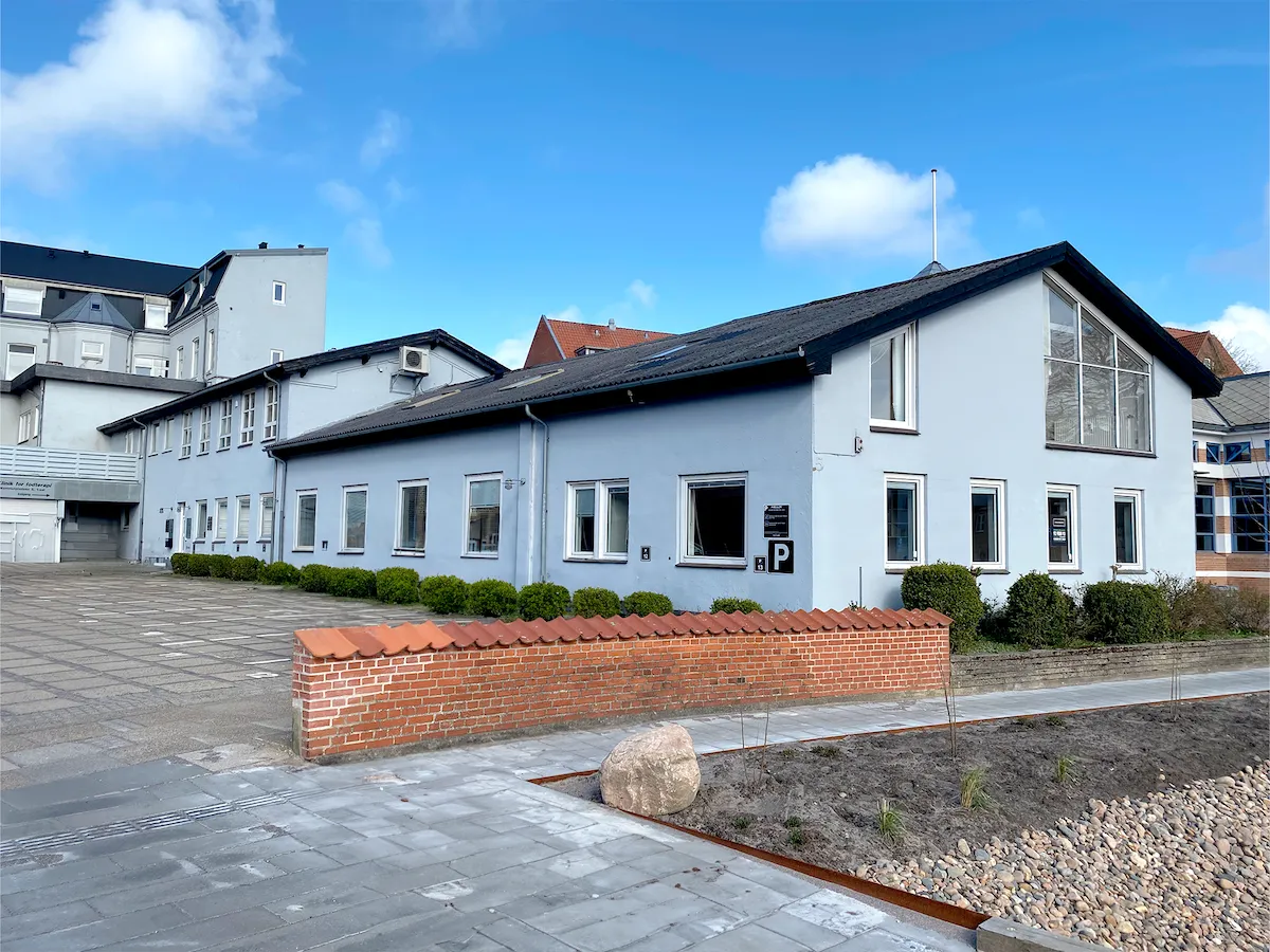 Moderne bygning med glasvinduer og sort tag på Mammutpladsen 5, 9800 Hjørring. Murstensvæg og grønne buske udenfor. Parkeringsskilt på facaden.