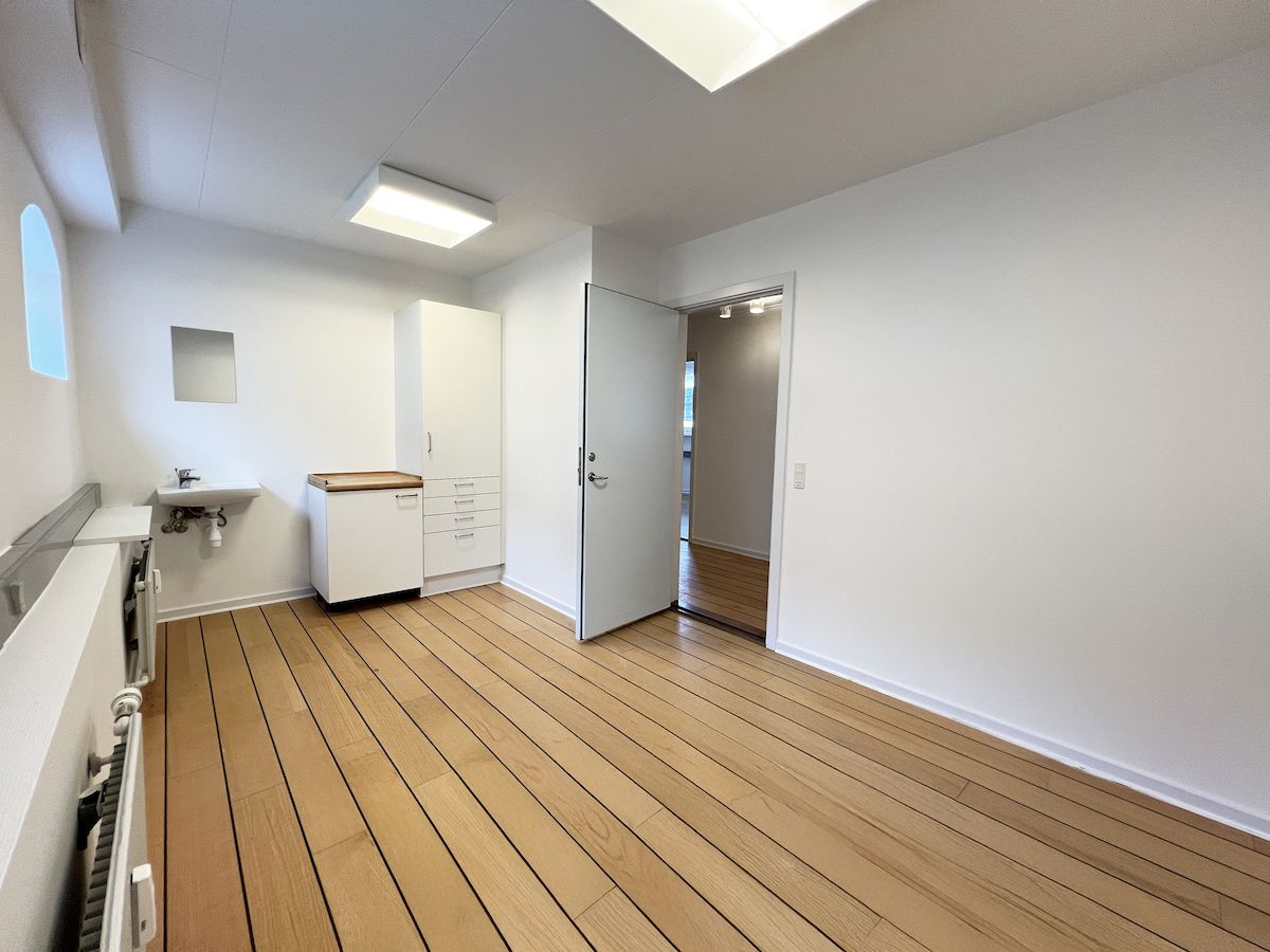 En ren og lys værelse med højt til loftet og hvide vægge. Værelset har trægulv og et moderne badeværelse med hvidt toilet og lysegrå væg.