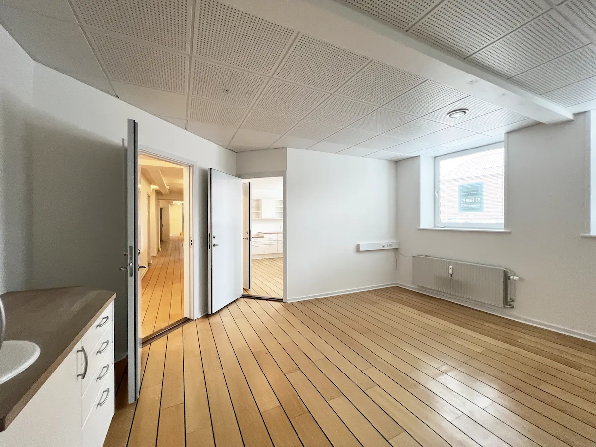 En stor, tom stue med lyse, trægulve, hvide vægge og åbne døre og vinduer. Et træbord med hvide skupe og en grå radiator mod væggen