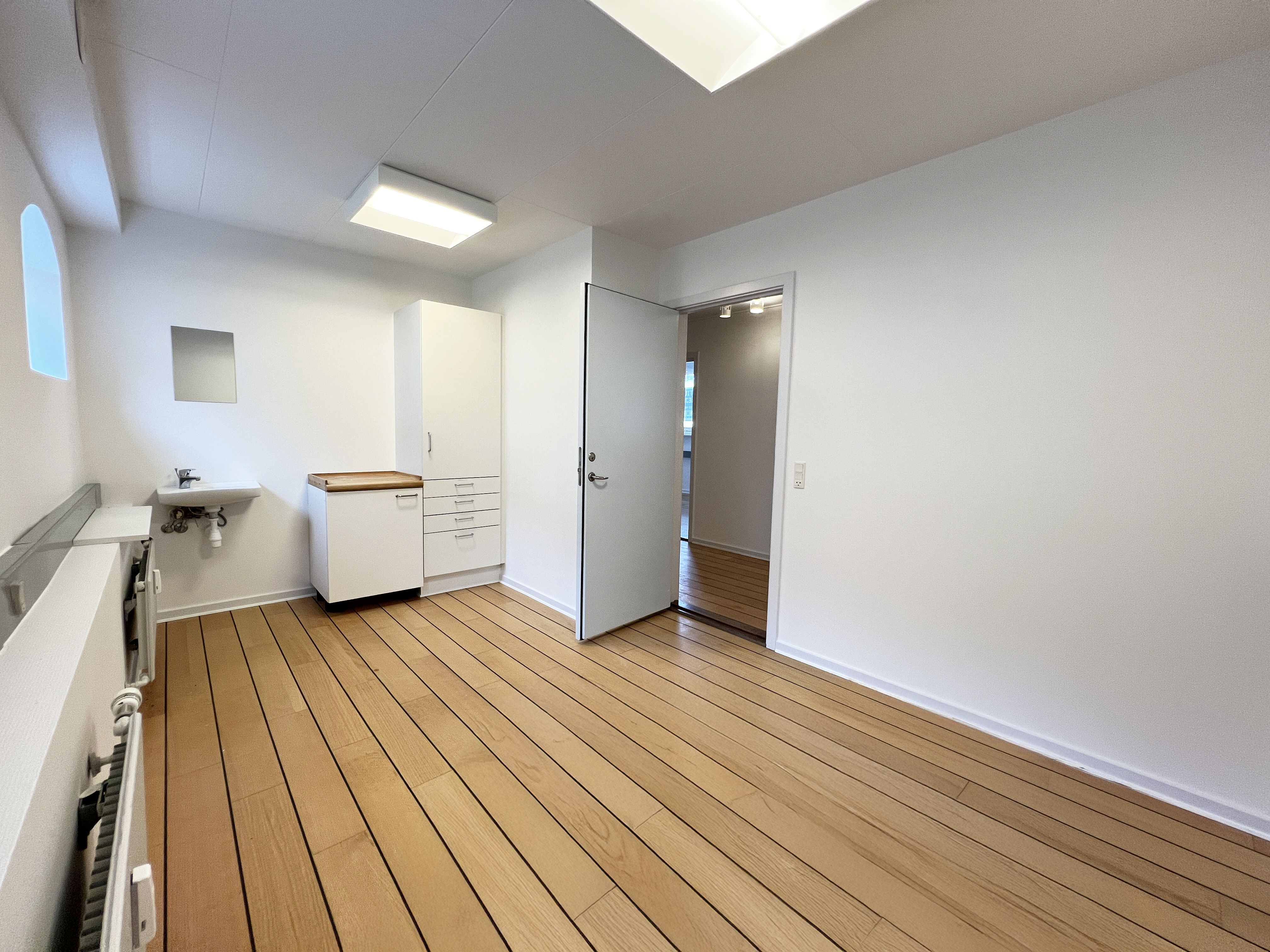 Foto af en rummelig klinik med hvidt loft og lysegule gulvplanker. Der er en åben dør og et lille skab med håndvask. Der er en træstolpe og et radiatorpanel på den ene side