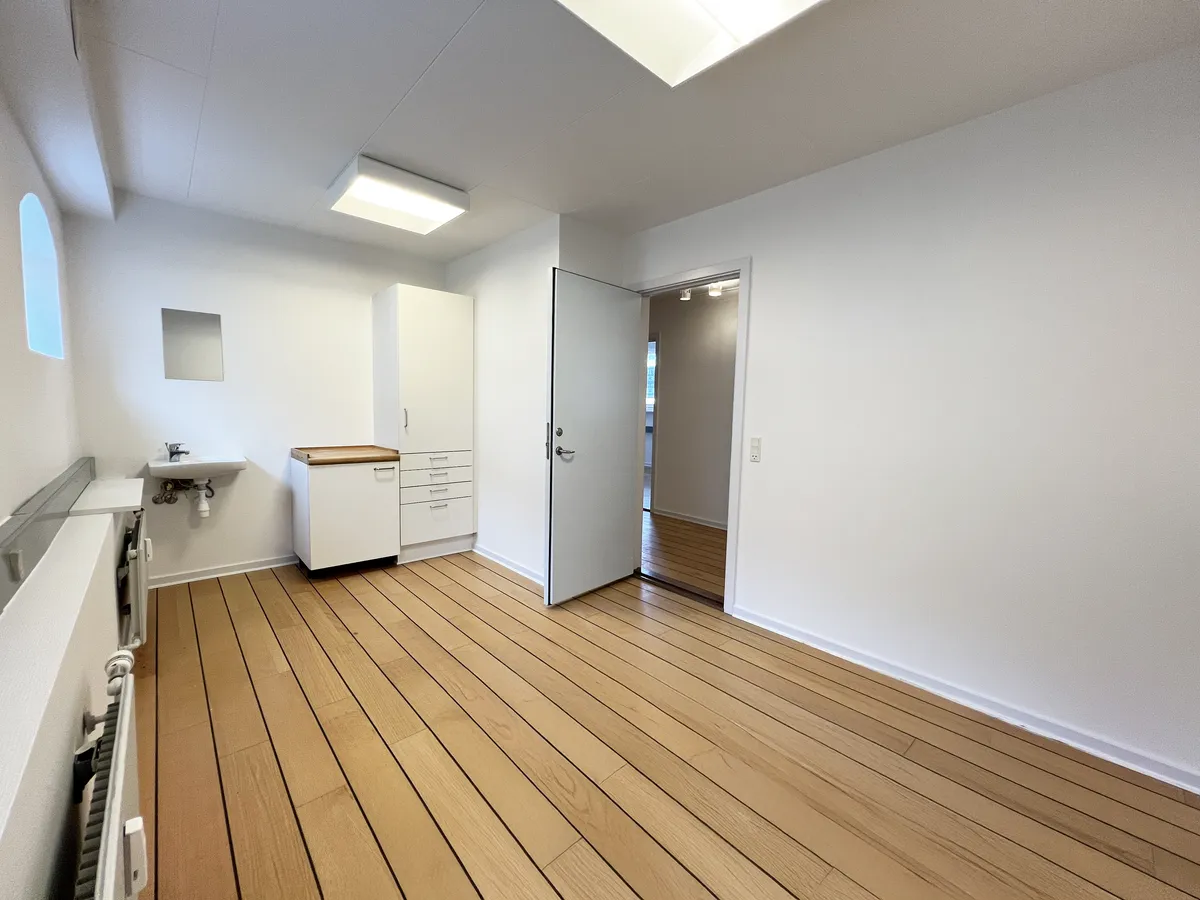 Foto af en rummelig klinik med hvidt loft og lysegule gulvplanker. Der er en åben dør og et lille skab med håndvask. Der er en træstolpe og et radiatorpanel på den ene side
