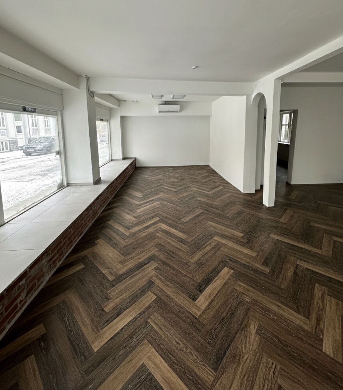 Moderne butikslokale med blanke hvide vægge og trægulv i bjælke og bjælke mønster, tæt ved Reberbansgade i Aalborg. Vinduet giver udsigt til en bilparkeringsplads og bygninger