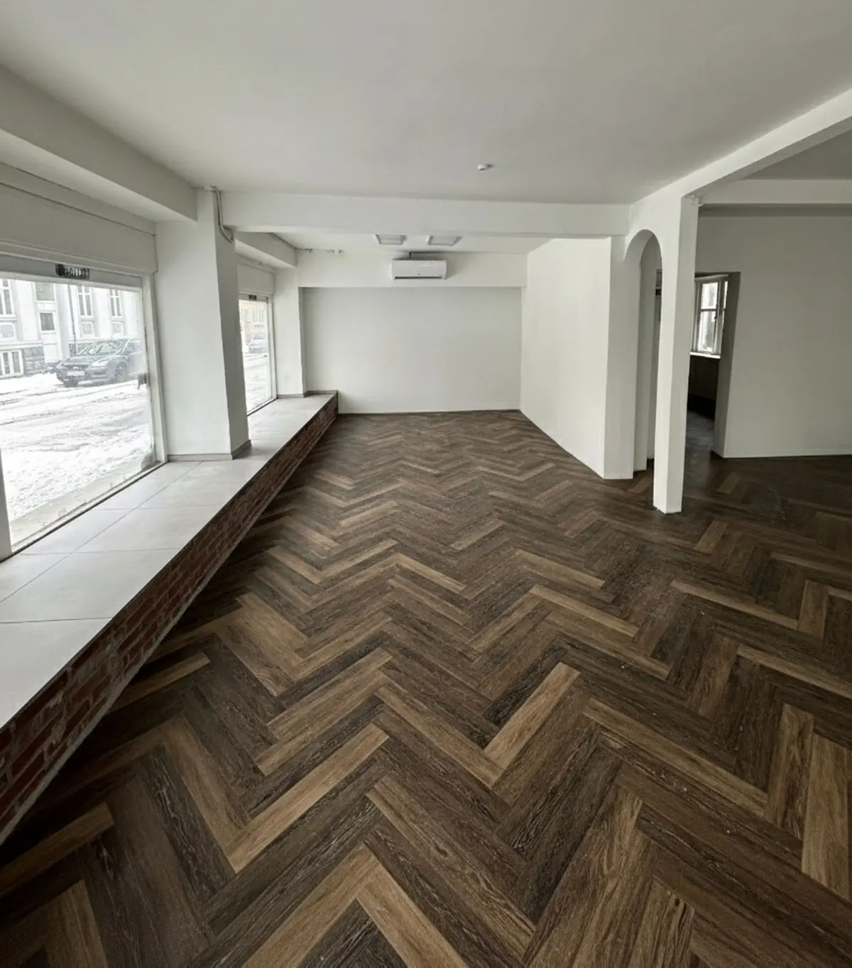 Moderne butikslokale med blanke hvide vægge og trægulv i bjælke og bjælke mønster, tæt ved Reberbansgade i Aalborg. Vinduet giver udsigt til en bilparkeringsplads og bygninger