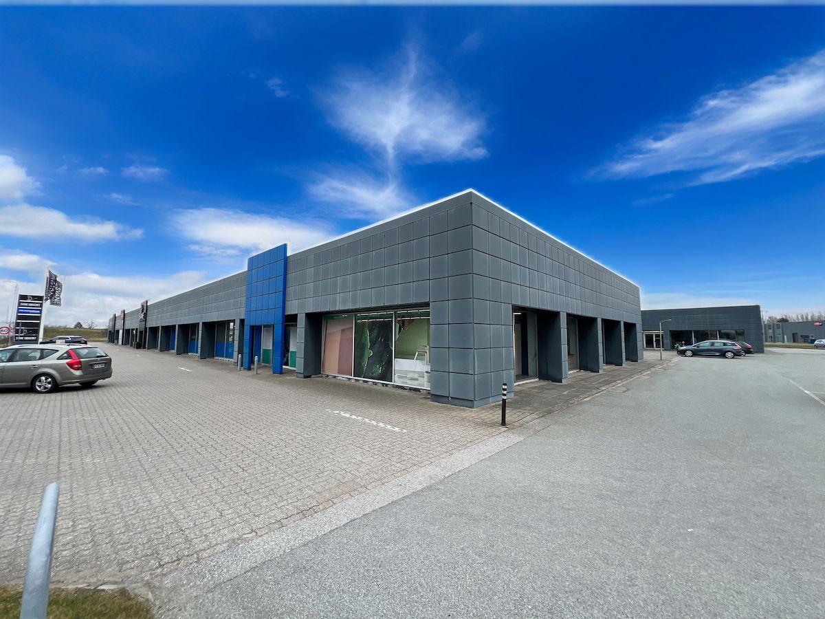 Moderne butiksbygning med glasvinduer og store eksponeringsflader, parkeringsplads og biler parkeret i forgrunden og en klar himmel med skyer i baggrunden