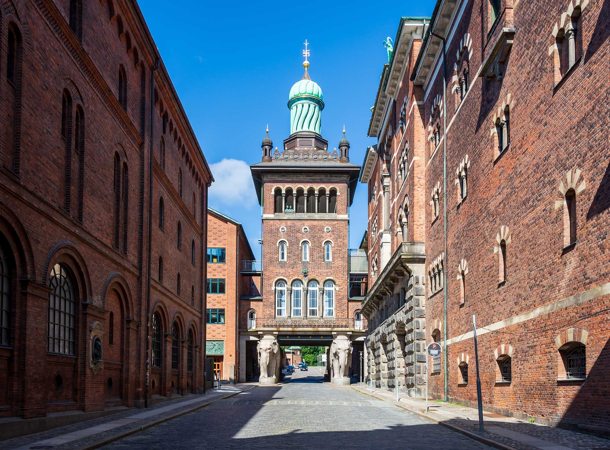 Rådgivende kontorer i hjertet af Carlsberg Byen med en stor indgang og to træstatuer. Murstensbygninger med mange vinduer og en lille grøn park.