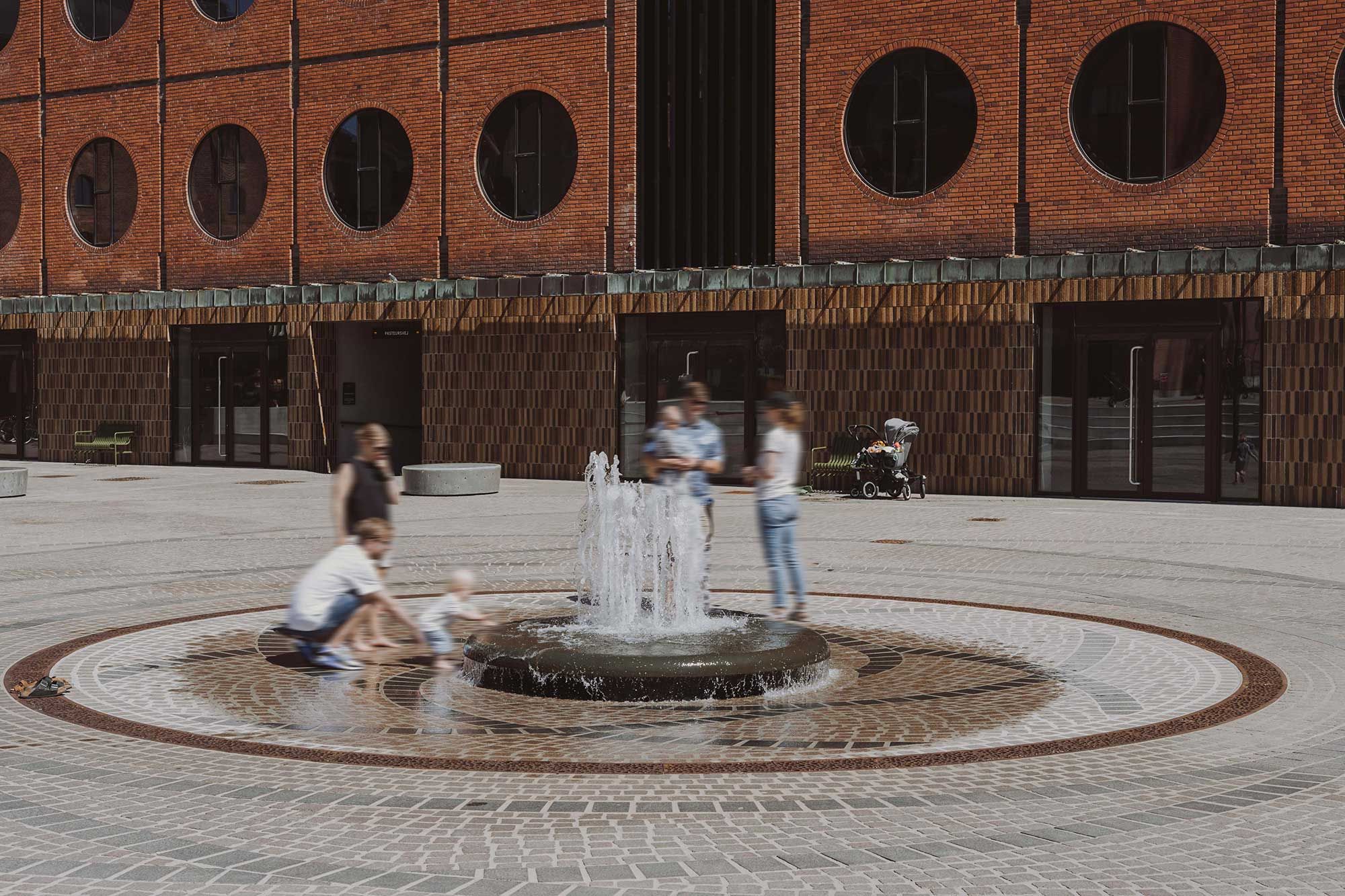 Mennesker og børn nyder vandspringet i gården. Bygningen med runde ruder og brun mursten er i baggrunden. Alle har skummet på skoene.
